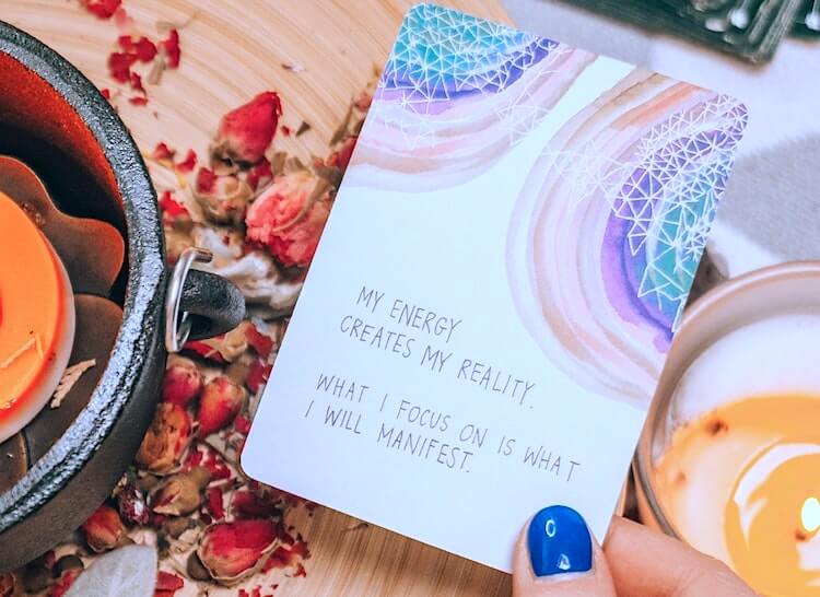 'Energy creates reality' manifestation quotes, with dried petals and candle in the background.