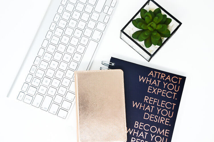A notebook with 'Attract what you expect' manifestation quote on its cover, beside a green succulent, a gold notebook, and a keyboard.