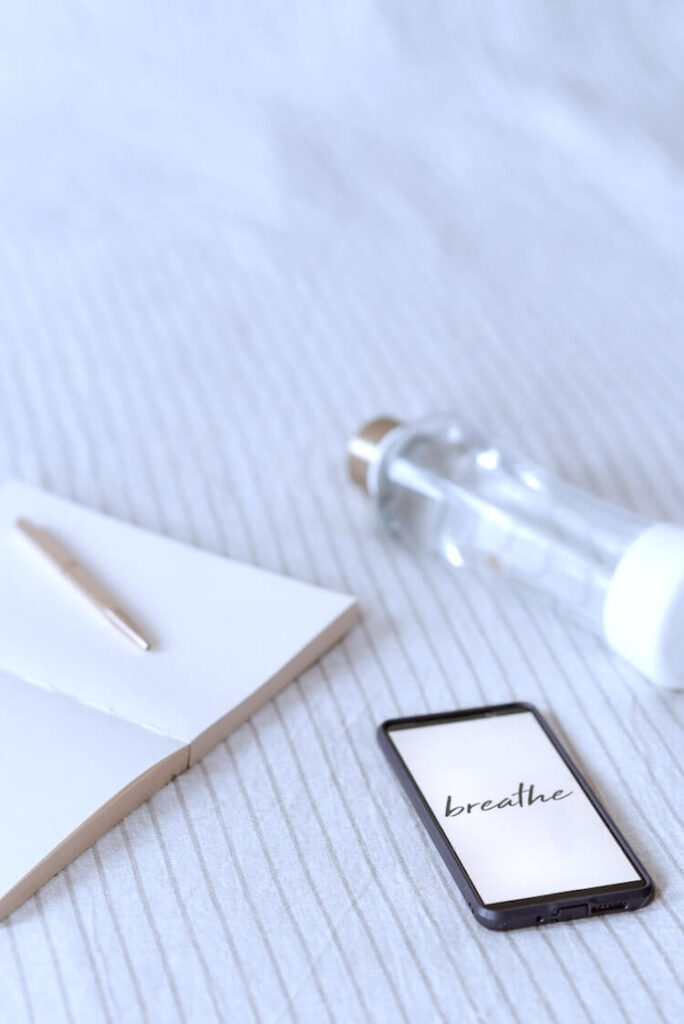 A mobile phone on a bed, with the word 'breathe' on the screen, beside a notebook and pen.