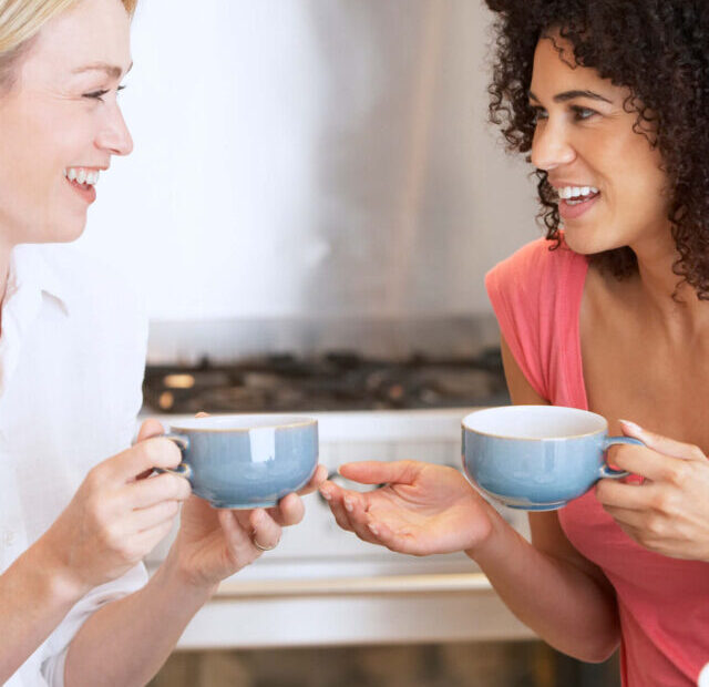 5/1 Human Design profile - Heretic Investigator | Two women sit beside each other with hot drinks in blue cups. One has shoulder length blonde hair and wears a white shirt, and the other has an afro and wears a coral coloured t-shirt.
