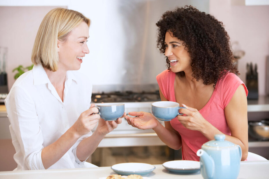 5/1 Human Design profile - Heretic Investigator | Two women sit beside each other with hot drinks in blue cups. One has shoulder length blonde hair and wears a white shirt, and the other has an afro and wears a coral coloured t-shirt.