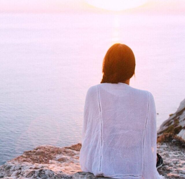 Incarnation cross Human Design | A woman sits on a rocky precipice overlooking the ocean. She wears a thin white shirt and has her back to the camera.