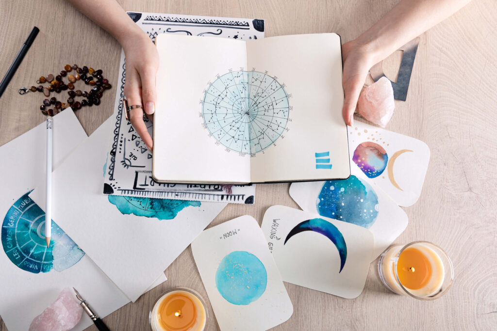 North Node 8th house astrology | An astrologist holds a watercolour painting of a map of the constellations in a notebook. There are also watercolour paintings on the desk of the moon in different phases, as well as a birth chart.
