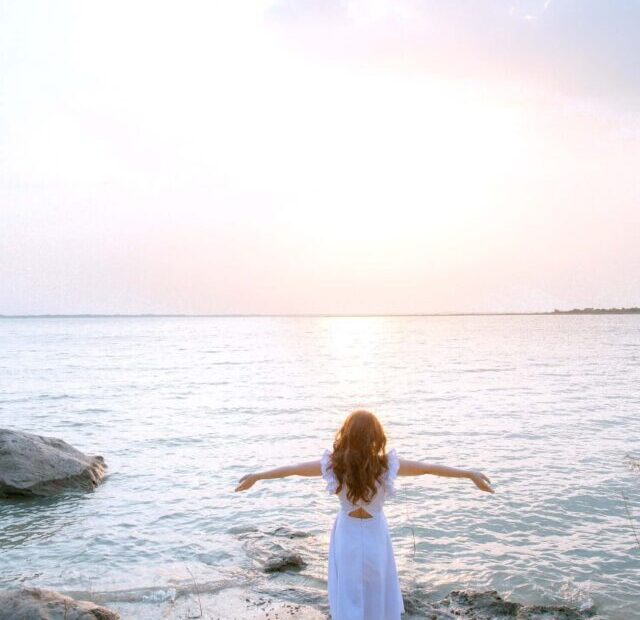 Human Design deconditioning | A woman in a white dress stands on the shore at dusk, looking out to sea, with outstretched arms in a freedom pose.