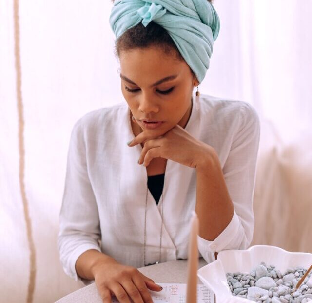 Human Design calculator reading | A woman with a mint green scarf tied around her head sits at a table for a Human Design reading. She wears a white shirt and sits with her chin resting on one hand and the other resting on a Human Design chart.